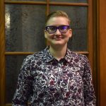 Bust shot of young adult girl with glasses, short blonde hair and in a button down flowered print shirt in reds and blacks with a collar. She is smiling and standing in front of a frosted pane of glass with oak framing.