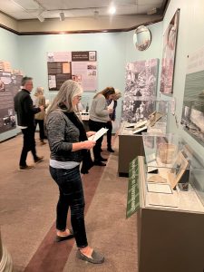 A woman standing next to an exhibit case displaying artifacts and archival materials. She is holding a sheet of paper. She is looking down at the paper. In the background, there are a couple of other people looking at other display cases. Attendees are solving riddles in the CSPM's "Evidence: Finding the Facts about General William Jackson Palmer" exhibit.