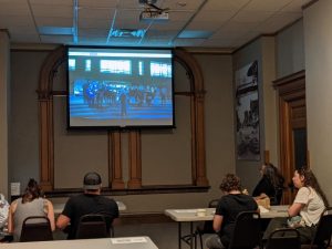 Four people sit at tables inside the Colorado Springs Pioneers Museum's Henderson room and watch a video on a screen at the front of the room.
