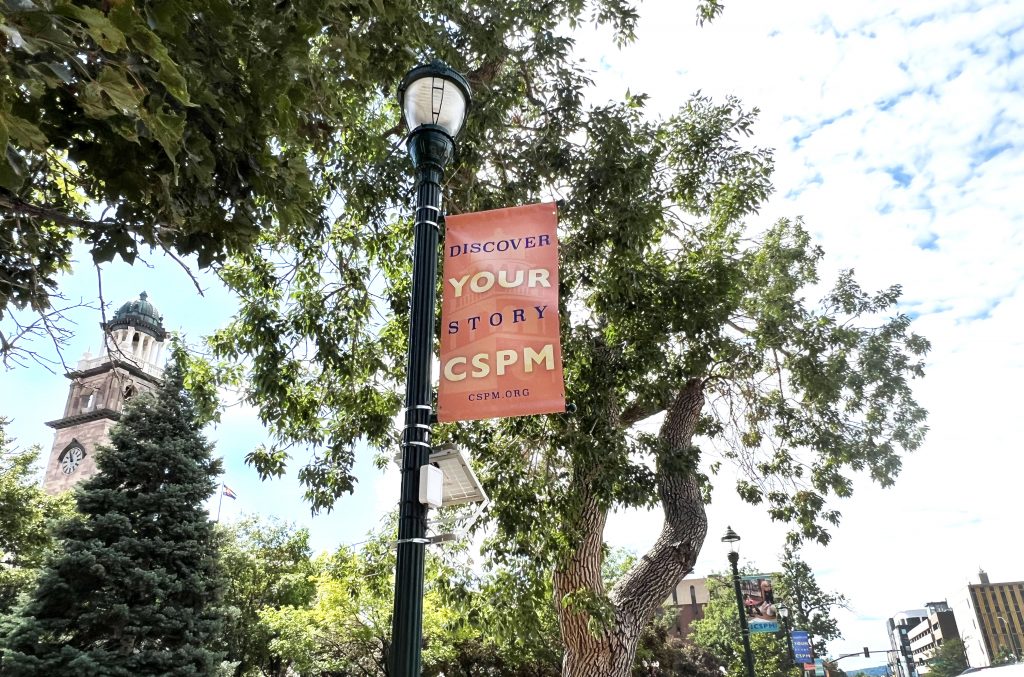 An orange banner that reads "Discover Your Story" and "CSPM" is mounted on a light pole. Subsequent banners can be seen along the street. The clock tower of the Colorado Springs Pioneers Museum is in the background.