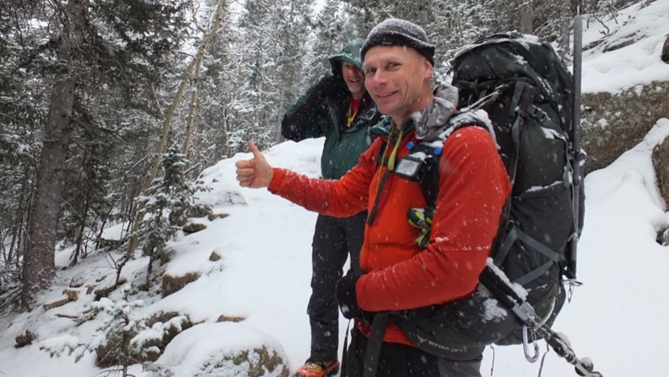 Lt. Gen. Christopher Coates is wearing a red sweat, one black glove and a hiking pack. He is giving the camera a thumbs up with his ungloved hand. There is another individual behind him.