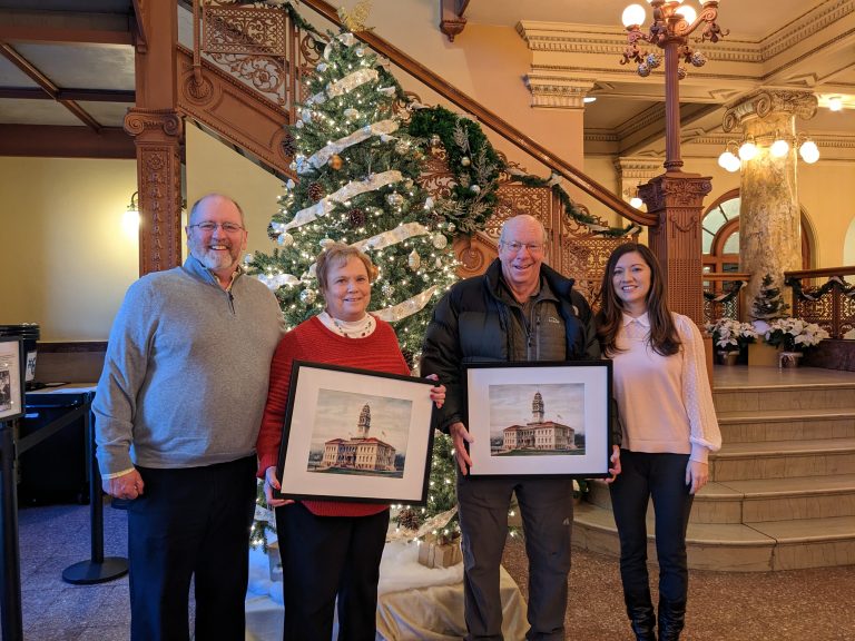 A group of four adults stand in front of a decorated Christmas tree and smile at the camera. A man and a woman in the middle each hold a framed illustration of the Colorado Springs Pioneers Museum.