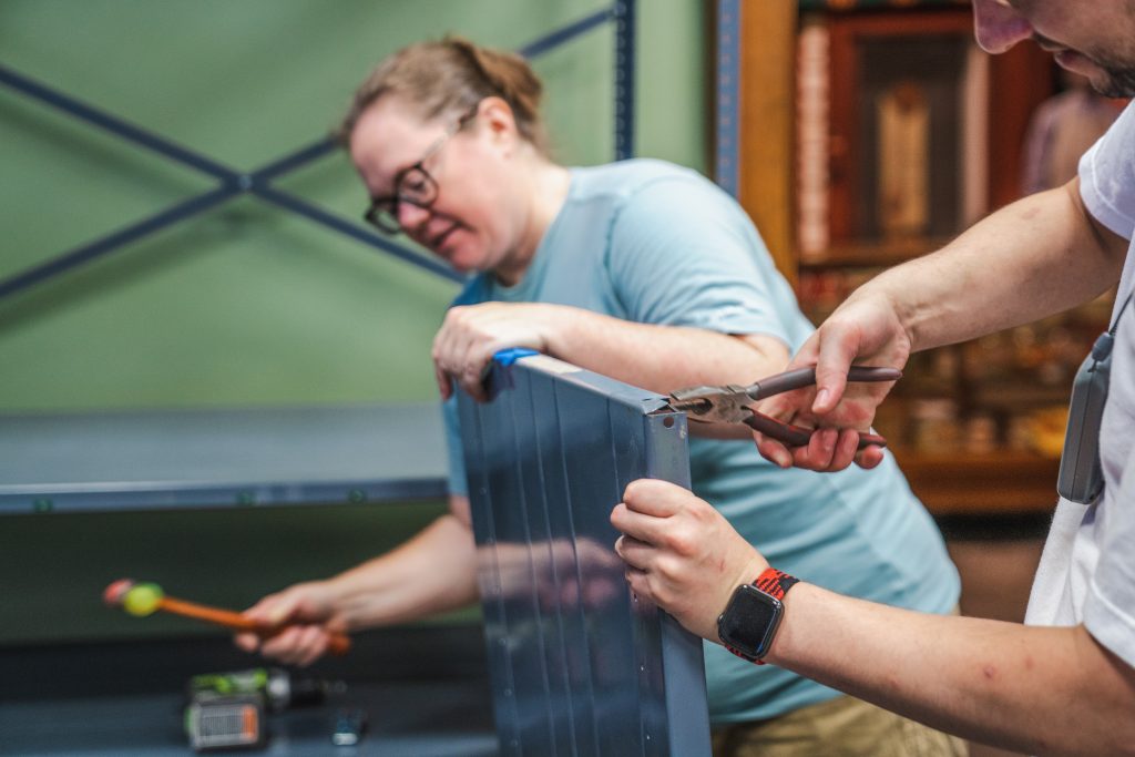 Holly Taylor is holding a hammer in her right hand and her left hand is holding the top of the shelf. She is about to hammer the lower part of the shelf. Alex is using pliers on the shelf's corner on the opposite side.