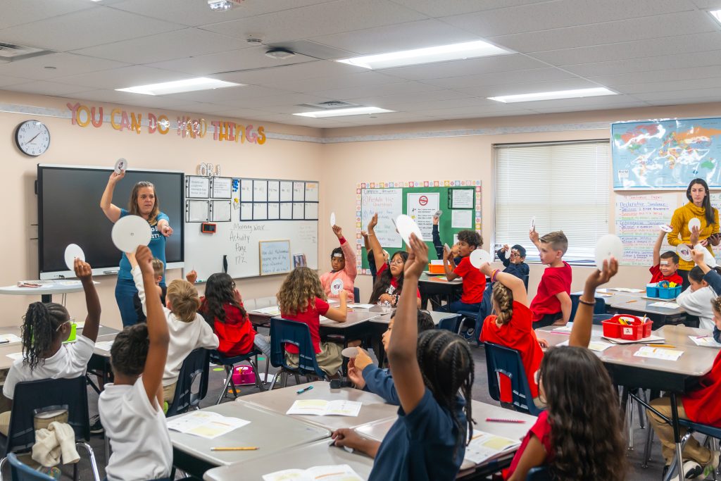 CSPM Education Outreach volunteers leading the History Detectives Outreach program for a group of students. The students are raising their hands with paper compasses as part of an activity.