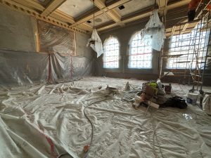 Division III Courtroom currently being rehabilitated into an exhibition gallery space. The room is empty except for plastic sheeting on the floor, the wood paneling and the chandeliers. There is also scaffolding on the right side of the room.