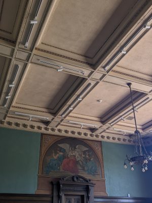 A photo focusing on the ceiling of the Division III Courtroom which includes the mural, corbels and chandeliors.