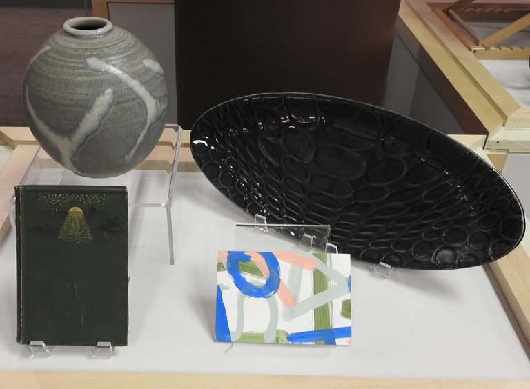Museum case; Black book with gold leaf detailing, pottery vase, handmade postcard, oblong handmade platter (left to right).