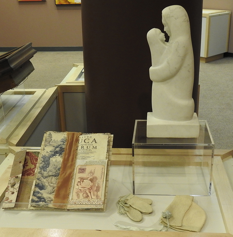 Museum case; Fabric scrap book, woolen mittens, sculpture of a mother and child, woolen hat (left to right).