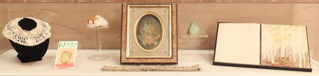 Museum case of collections items; Lace collar, homemade postcard, angel figurine, shadowbox floral pressing, lace strap, ceramic rabbit, and artist sketchbook (left to right).