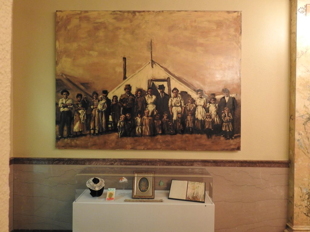 Above, painting of pioneer family gathered in front of an oversized tent; Below, museum case of collections objects.
