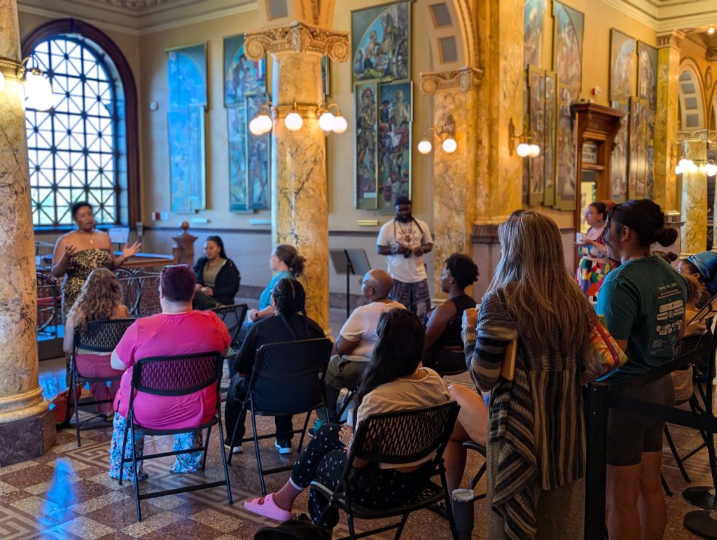 Program participants sit in chairs in the museum lobby while the program presenter, Ashley Cornelius, speaks to them.