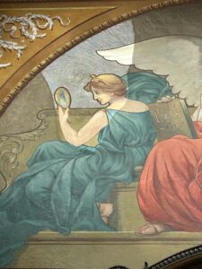Close up image of the Division III Courtroom mural focusing on the figure holding a mirror. You can see the difference between the treated and untreated areas of the mural. A section is brighter compared to the rest of the mural.